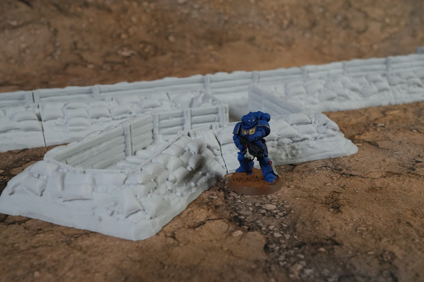 Detaillierter weißer Schützengraben mit Wände und Bodenstruktur neben blauer Miniaturfigur auf braunem Untergrund.
