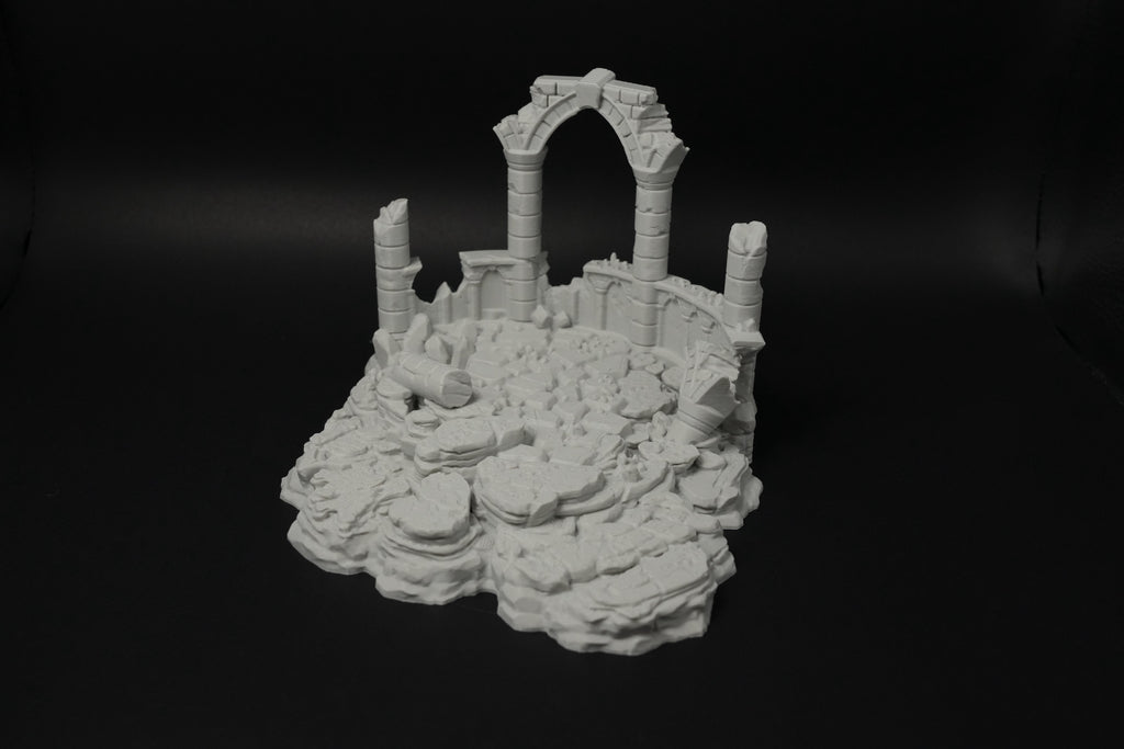Graue Miniatur antiker verwitterter Ruinen mit Säulen und einem Bogen auf felsigem Grund.