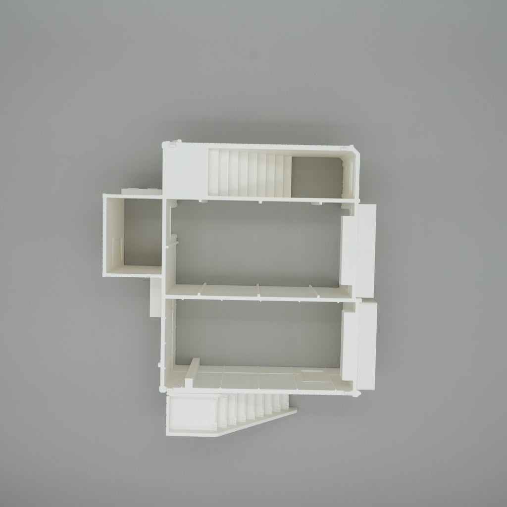 Weißes Modell eines großen Wohnhaus-Grundrisses mit Treppenhaus und mehreren Räumen auf grauem Hintergrund.