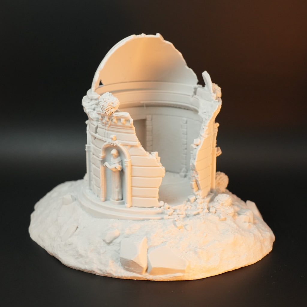 Weißes, zerfallenes Rundgrab mit verzierten Mauerresten und zertrümmertem Kuppeldach auf unebenem Sockel.