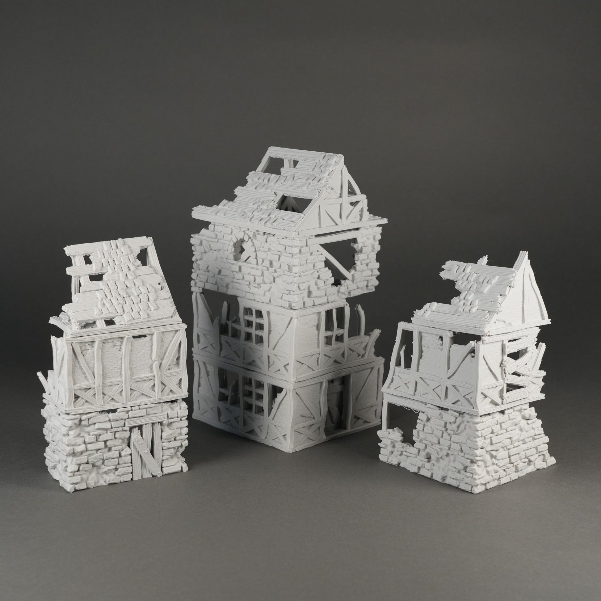 Drei graue Miniatur-Hausruinen mit Fachwerk und zerfallenen Dächern auf dunklem Hintergrund.