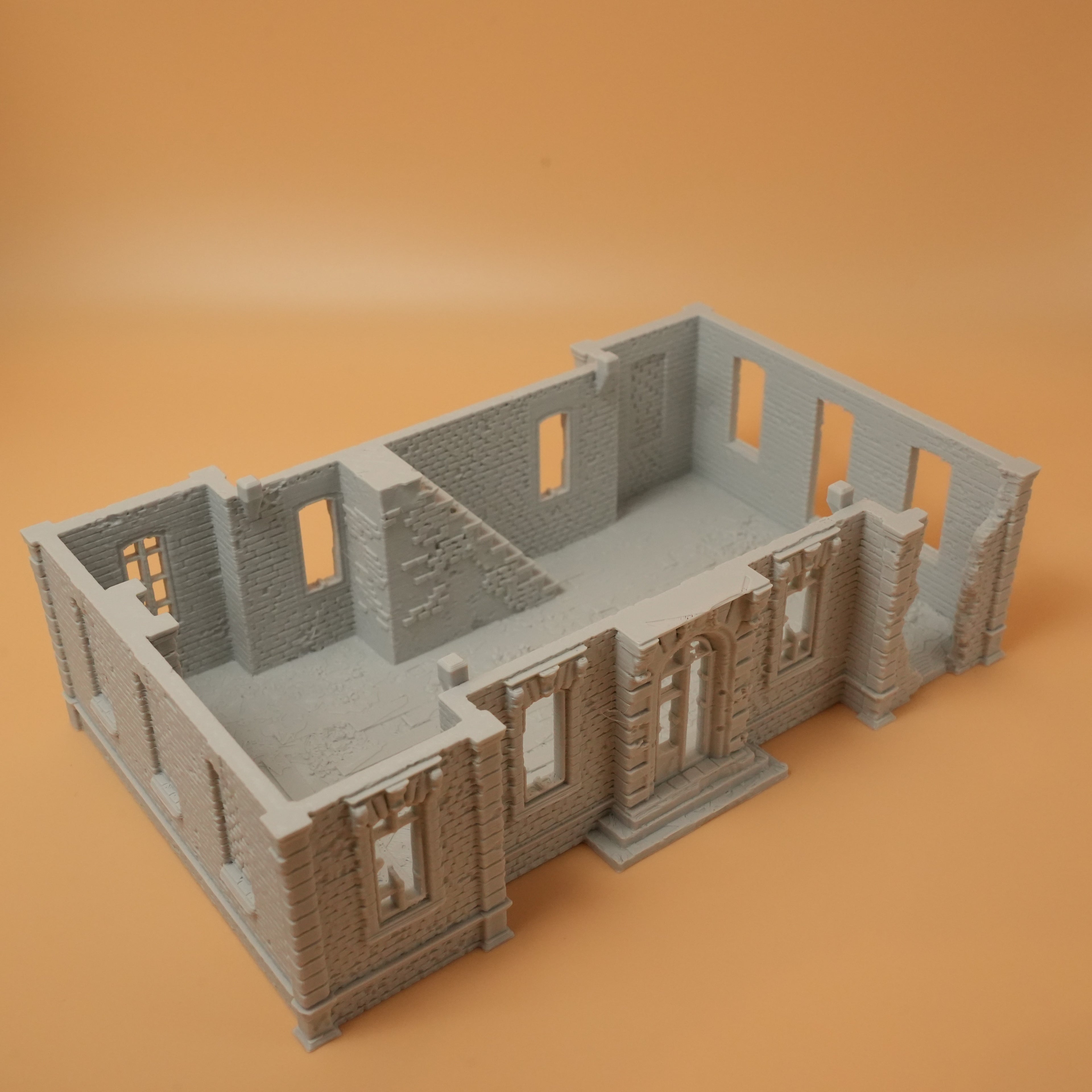 Graue Miniatur eines zerstörten Herrenhauses mit teils eingestürzten Wänden auf beigem Hintergrund.