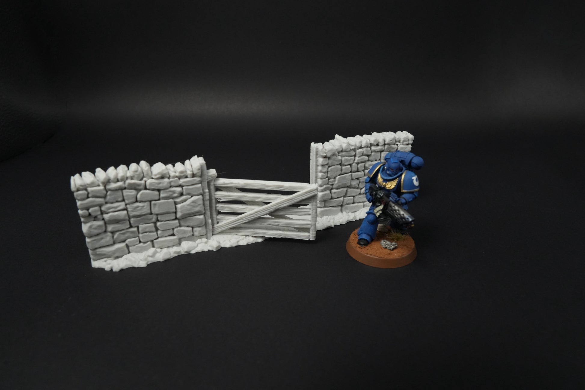 Hohe graue Steinmauern mit Holztor und Miniaturfigur daneben, schwarze Oberfläche.