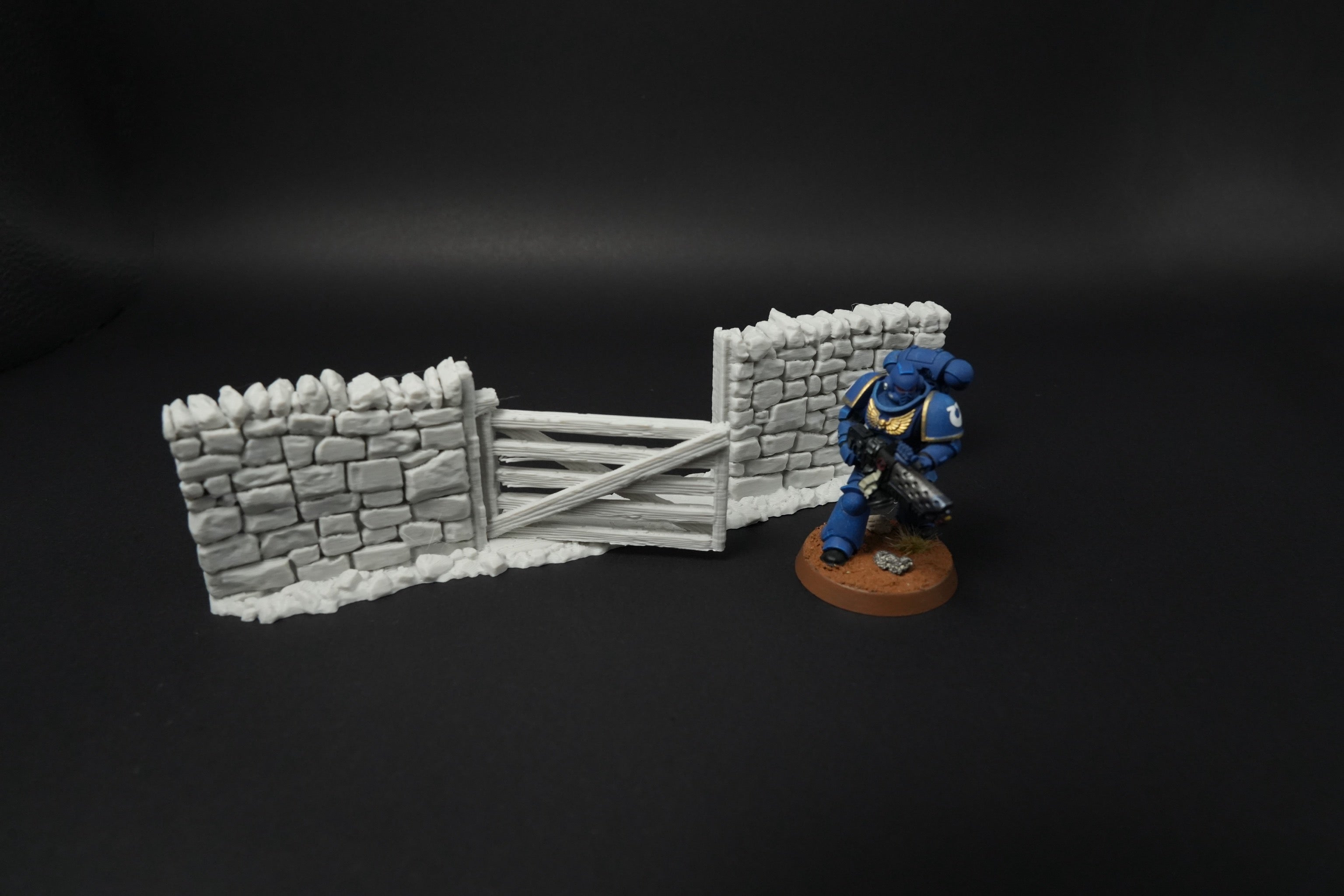 Hohe graue Steinmauern mit Holztor und Miniaturfigur daneben, schwarze Oberfläche.