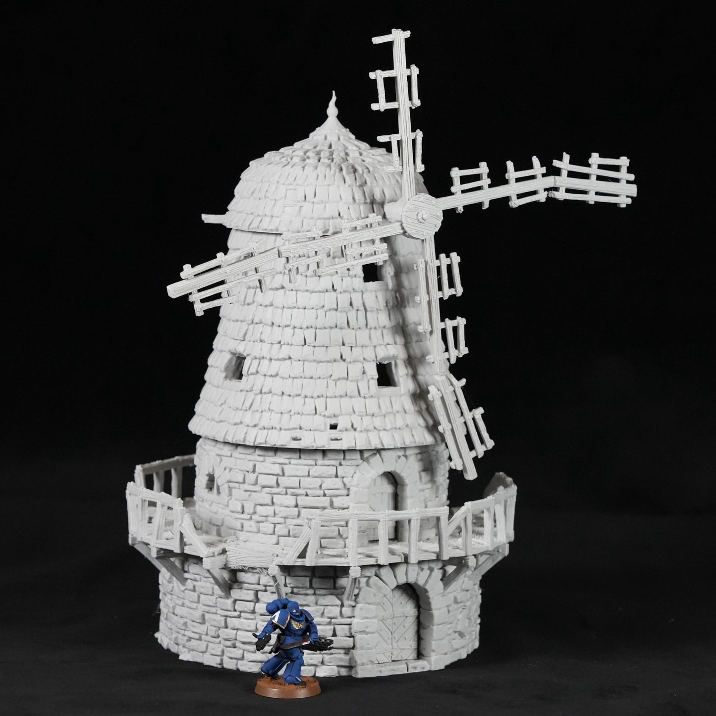Graue Windmühle aus Stein mit gebogenen Flügeln und Balkon, davor kleine blaue Figur.